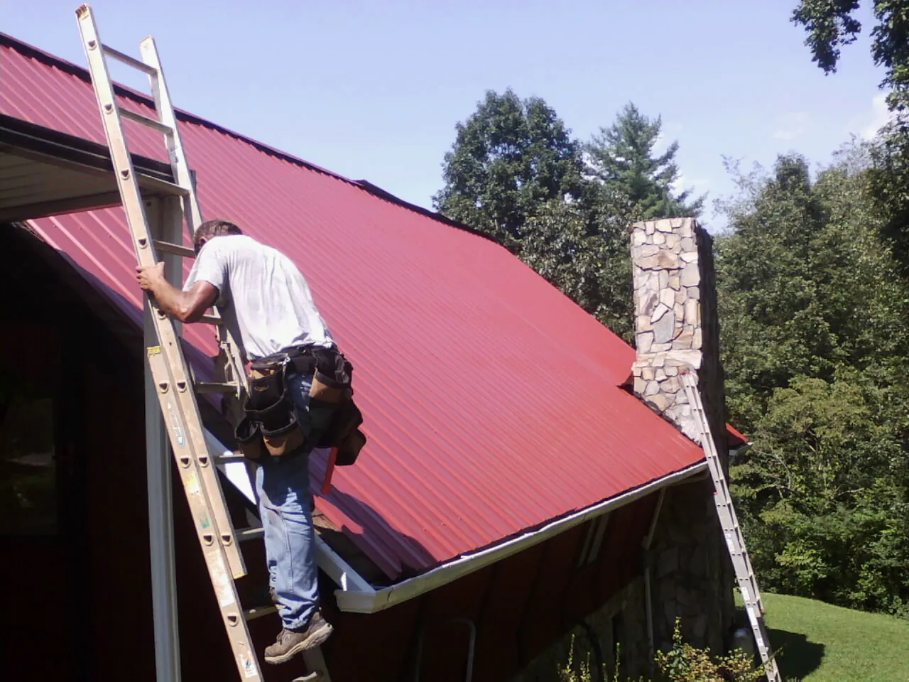 Roofer on a ladder beside a red metal roof and stone chimney — Soffit & Fascia Repair by Roofing Brothers in Camp Sherman